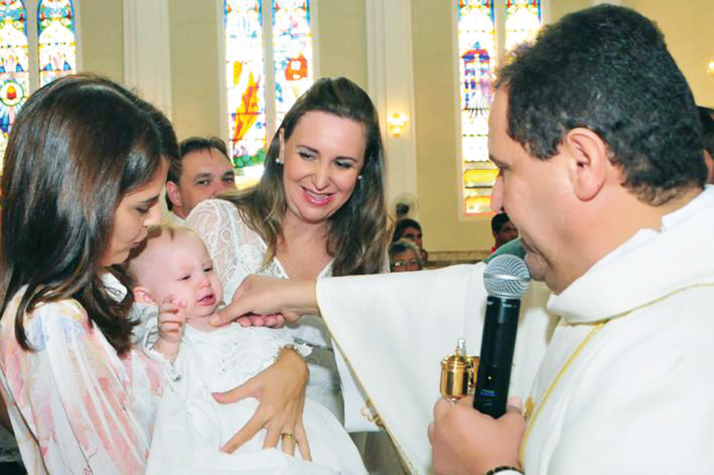 A pequena Joana, herdeira dos corujas papais Sandro Rajuna e Ana Paula Stefanelli em foto durante seu batizado, no colo da madrinha de consagração Luciana Garcia. Benção do pároco Gilmar