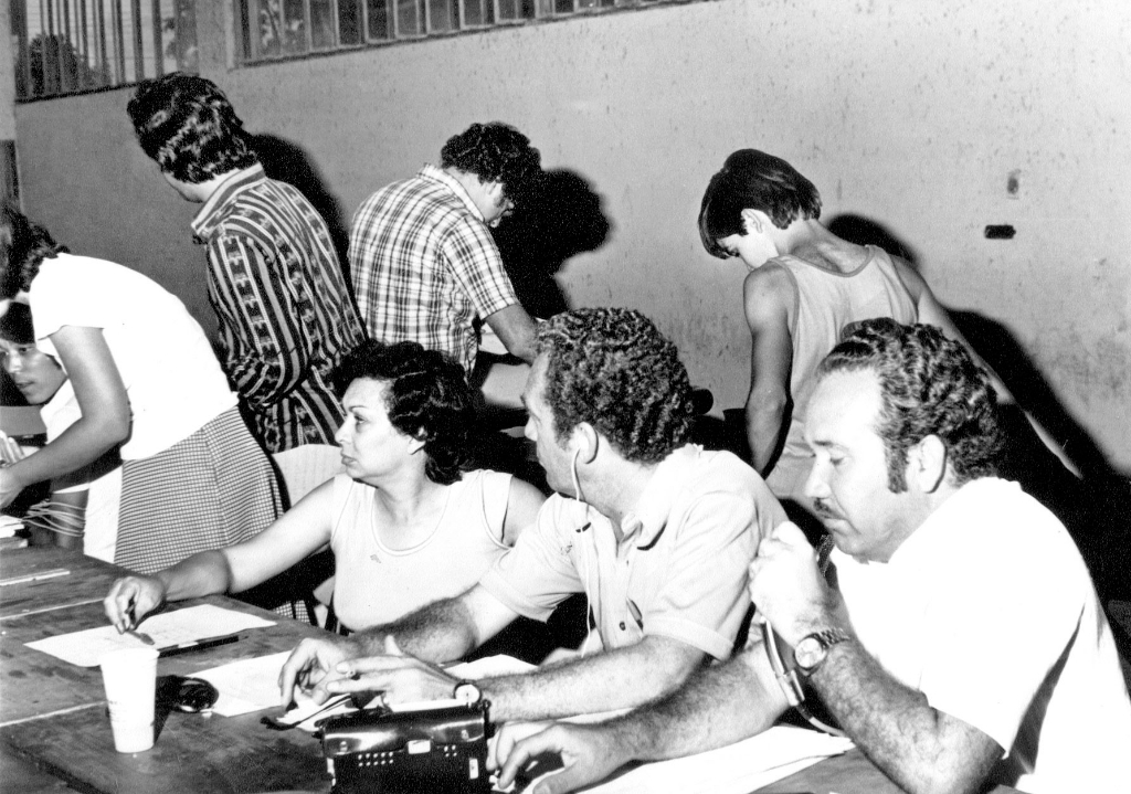 Em ação a equipe da Rádio Clube AM na cobertura da apuração dos votos na eleição de 1976. Naquele pleito eleitoral o dentista João Antonio Nucci elegeu-se prefeito pela primeira vez. O radialista Aguinaldo de Oliveira (à direita com o microfone) foi eleito o vereador mais votado com 768 votos da Arena Na esquerda, o técnico de som Shoyo e na mesa o casal de radialistas Luizz Rivioiro e Oneide Aparecida Lourenço Rivoiro.  Atrás, de costas, Edélcio Martins e Celso Fiorentino. A foto pertence ao álbum de Oliveira.